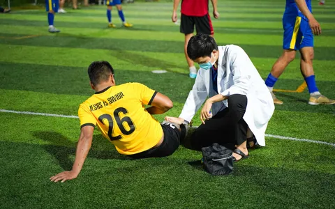 Atleta sentado no gramado segurando a parte posterior da coxa após uma lesão