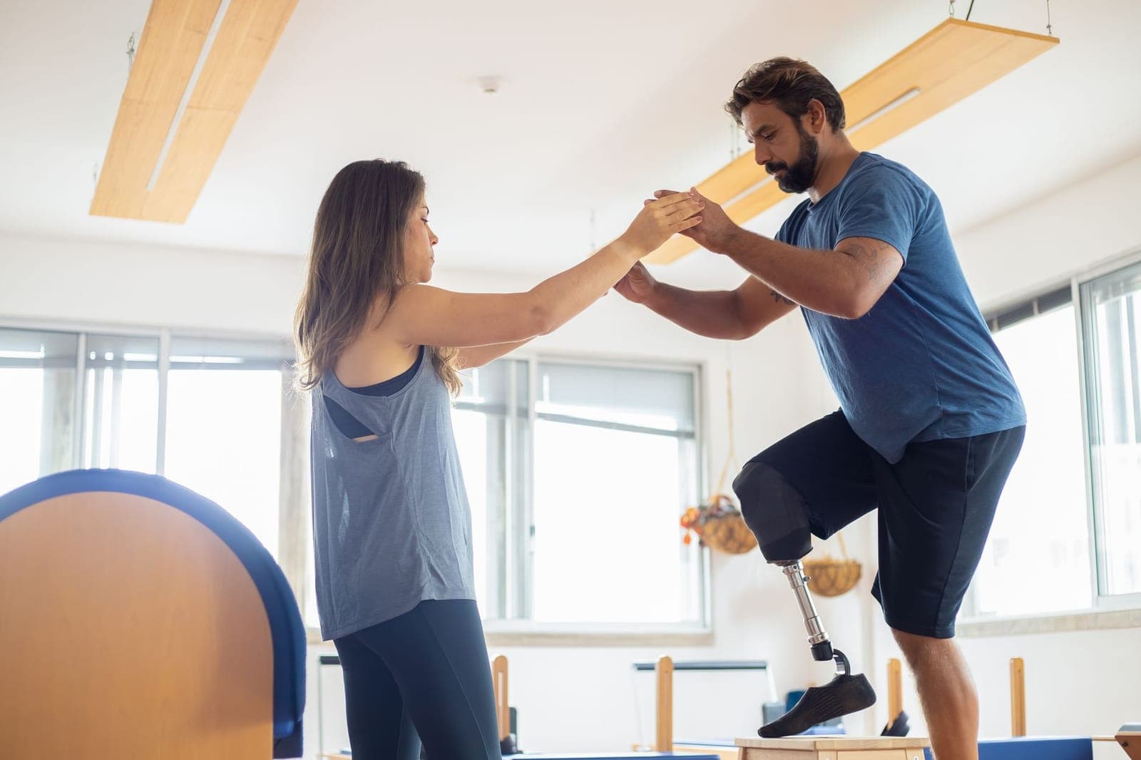 Fisioterapeuta segurando as mãos de um paciente com prótese de perna durante exercício de equilíbrio sobre um step